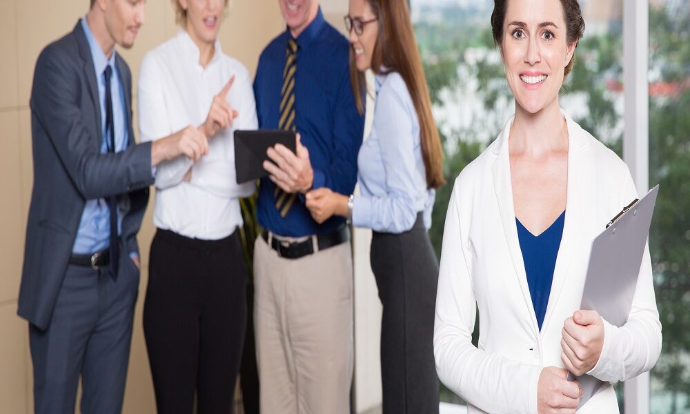 Portrait of happy young female executive manager standing in office, holding clipboard, smiling at camera. Her business team discussing project on tablet computer in background. Leadership concept